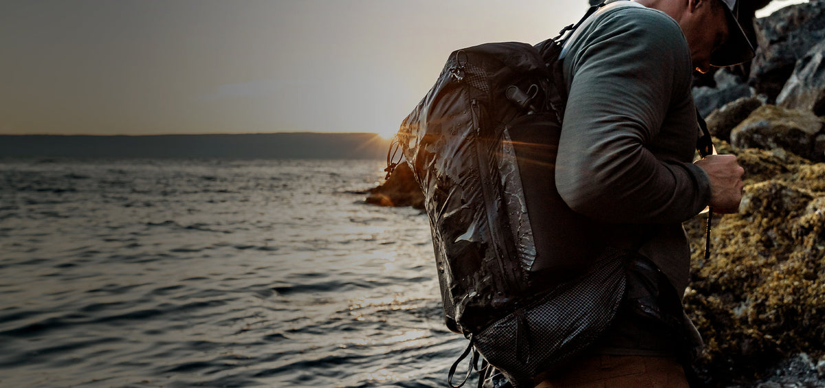 Man on rocky sunset shore, wearing a wet backpack