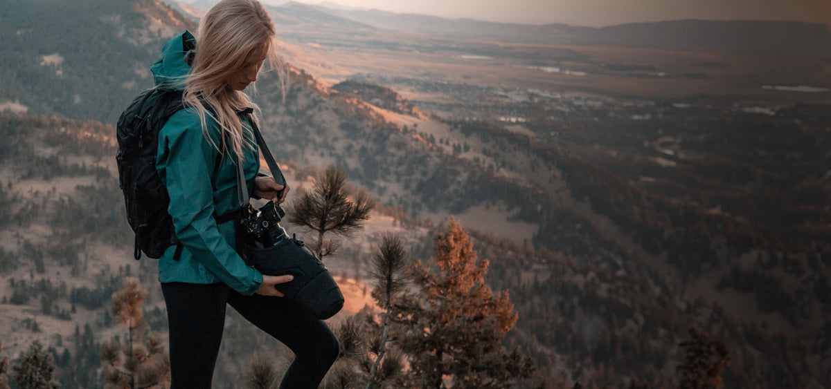 Woman in front of sunset mountains, pulling camera out of her camera baselayer