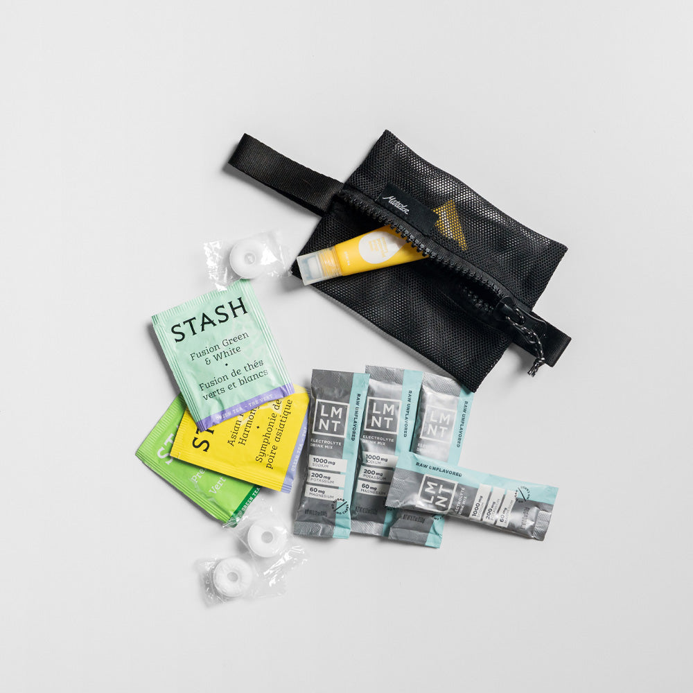 Small mesh bag on light gray studio background with items spilling out: teas, mints, LMT powder, lip gloss